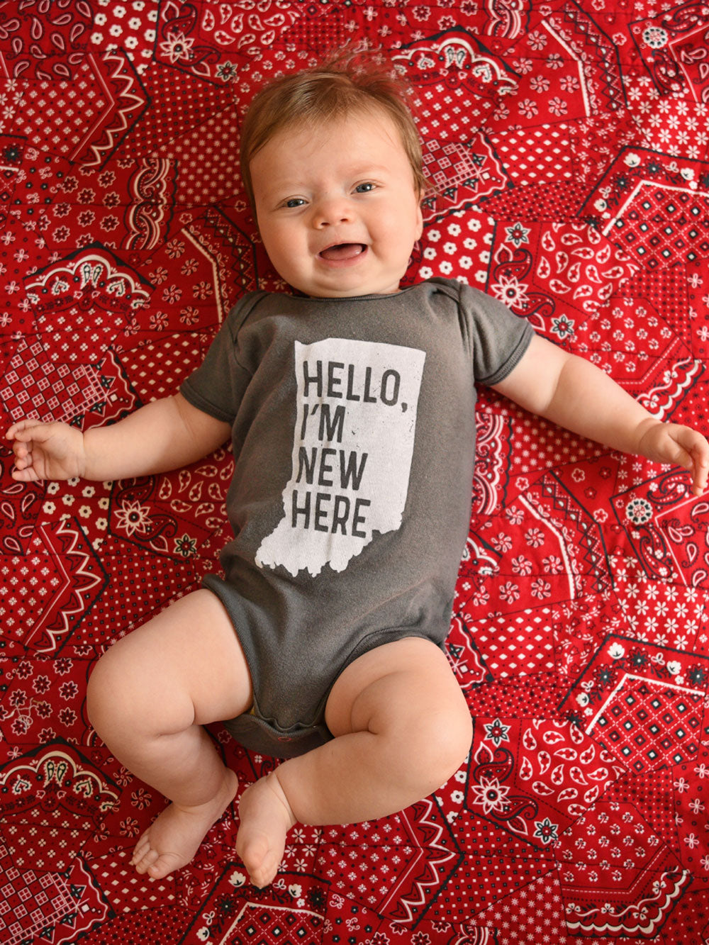 Hello, I'm New Here (Indiana) gray infant bodysuit on baby laying on red bandana quilt